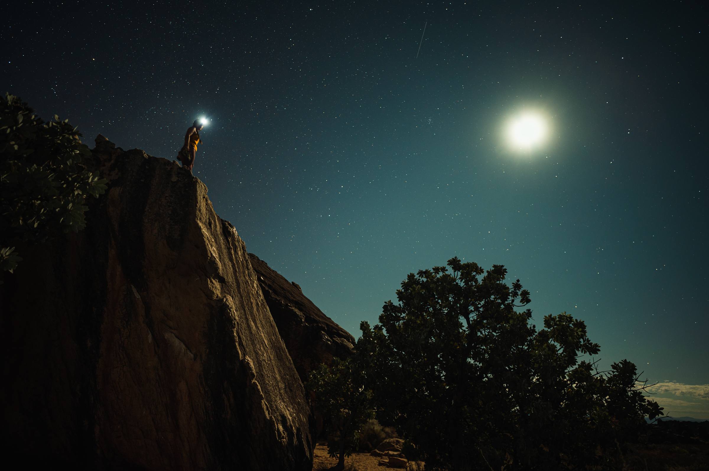A climber standing on the top of a Rocklands boulder by headlamp under a bright moon and stars