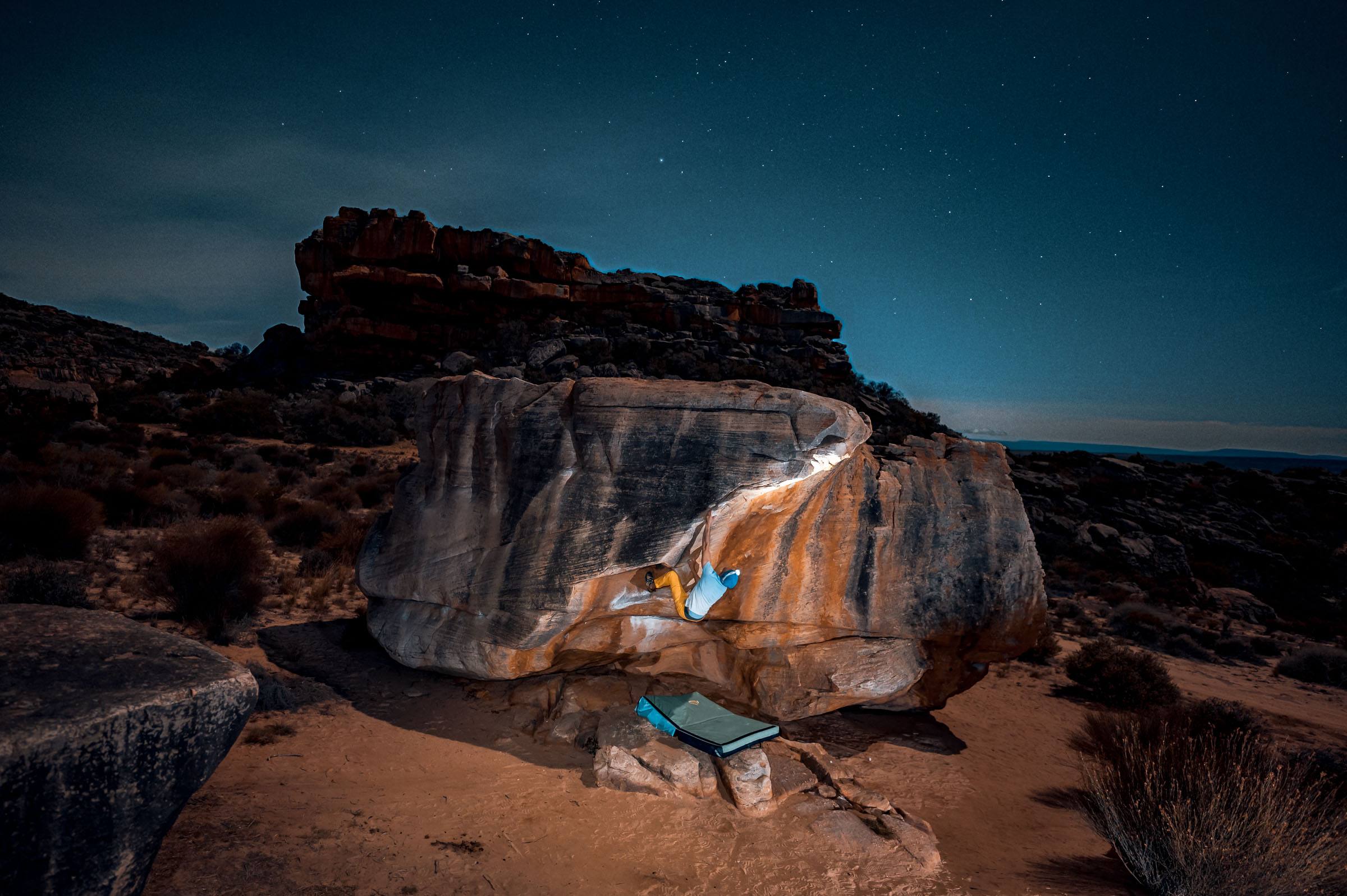 A lit sandstone boulder with a climber on it under a star-filled Rocklands night sky, a crash pad below