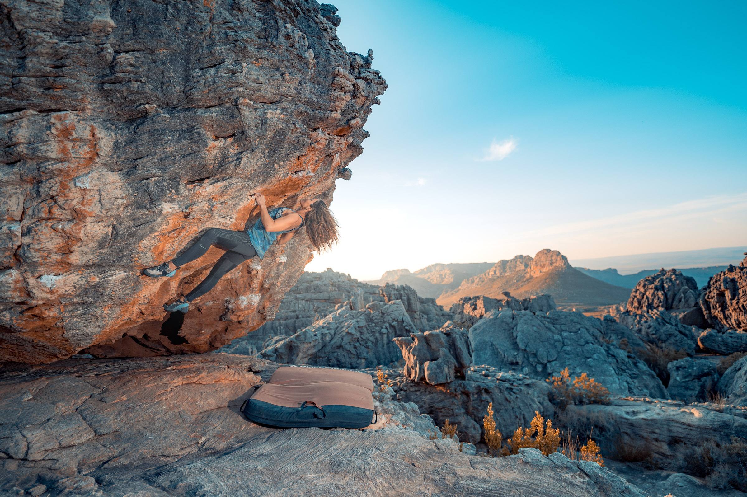 A boulderer cutting loose on an overhanging Rocklands boulder with the Cederberg peaks behind in clear winter light