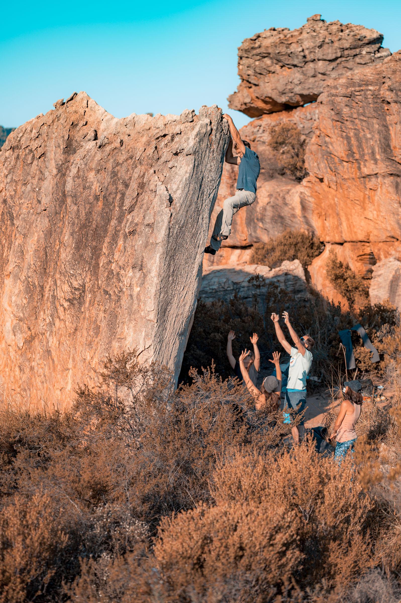 A climber topping out a tall Rocklands boulder while a group of spotters waits below with hands raised