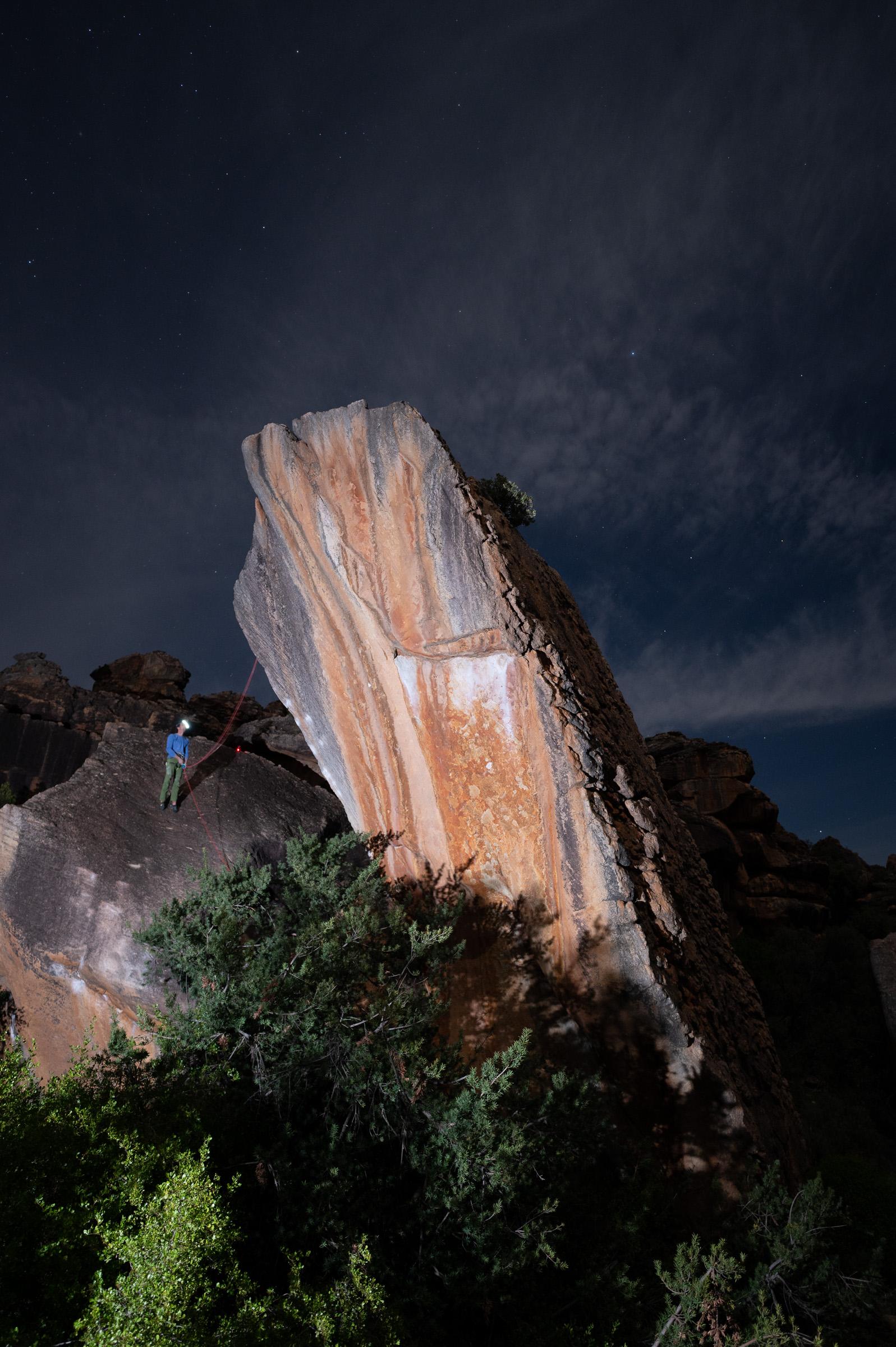 A tall, water-streaked sandstone boulder at Rocklands lit against the night sky, a climber small at its base