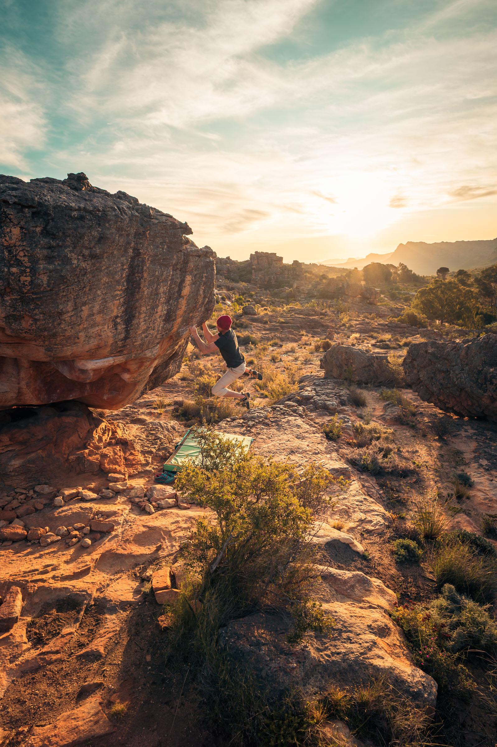 A boulderer pulling onto an overhanging sandstone boulder at Rocklands at sunset, a crash pad on the ground below