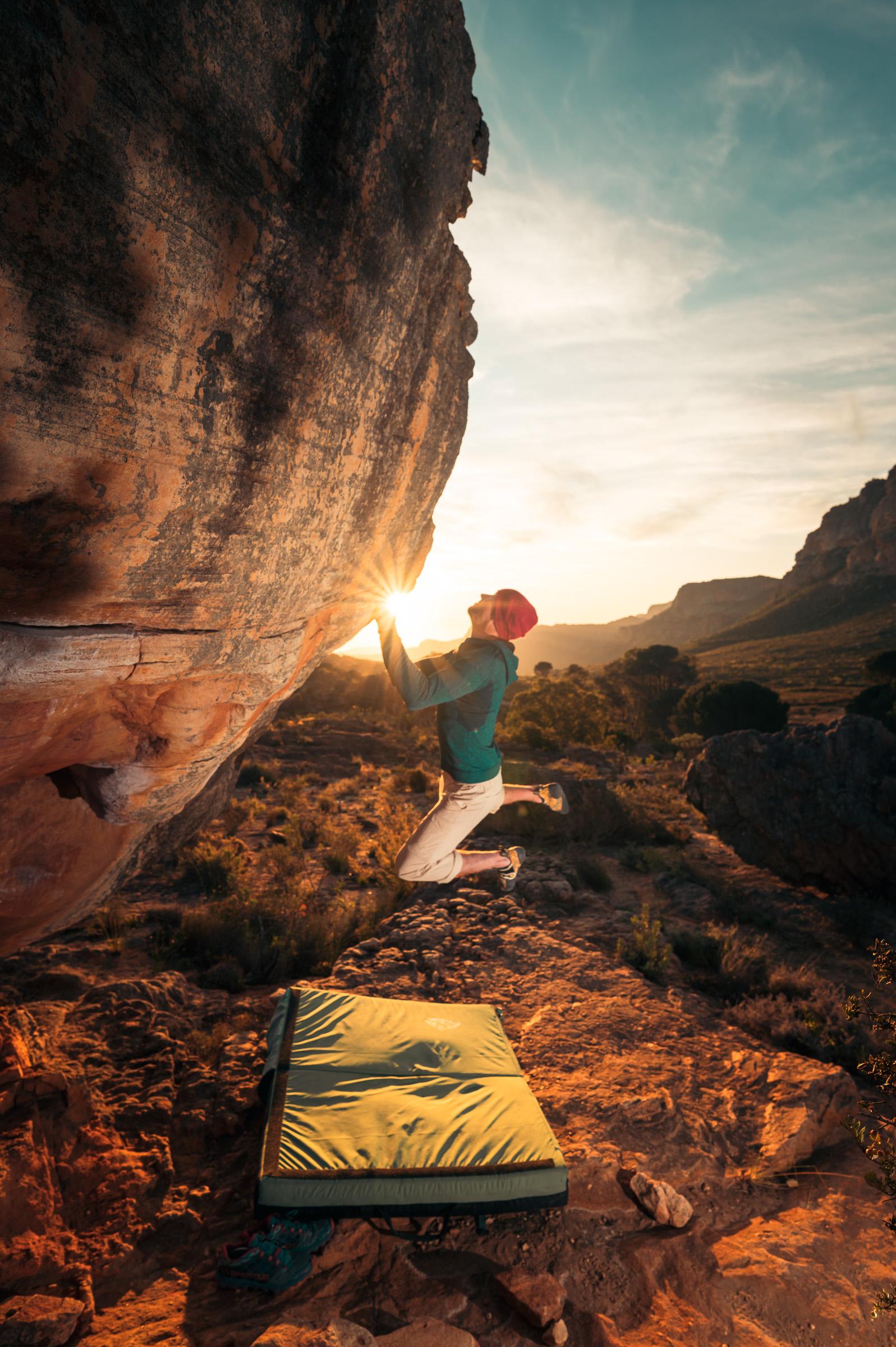 A climber reaching for the lip of an overhanging Rocklands prow as the sun sets behind the Cederberg mountains