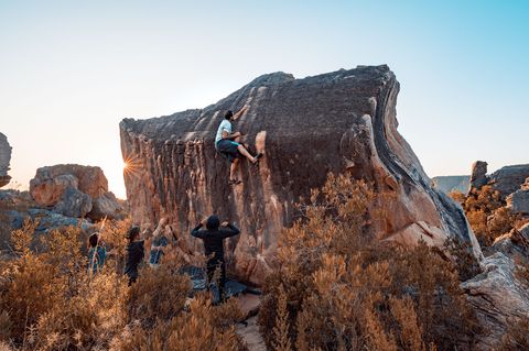 A boulderer topping out a large sandstone boulder at Rocklands with spotters below and the low sun behind